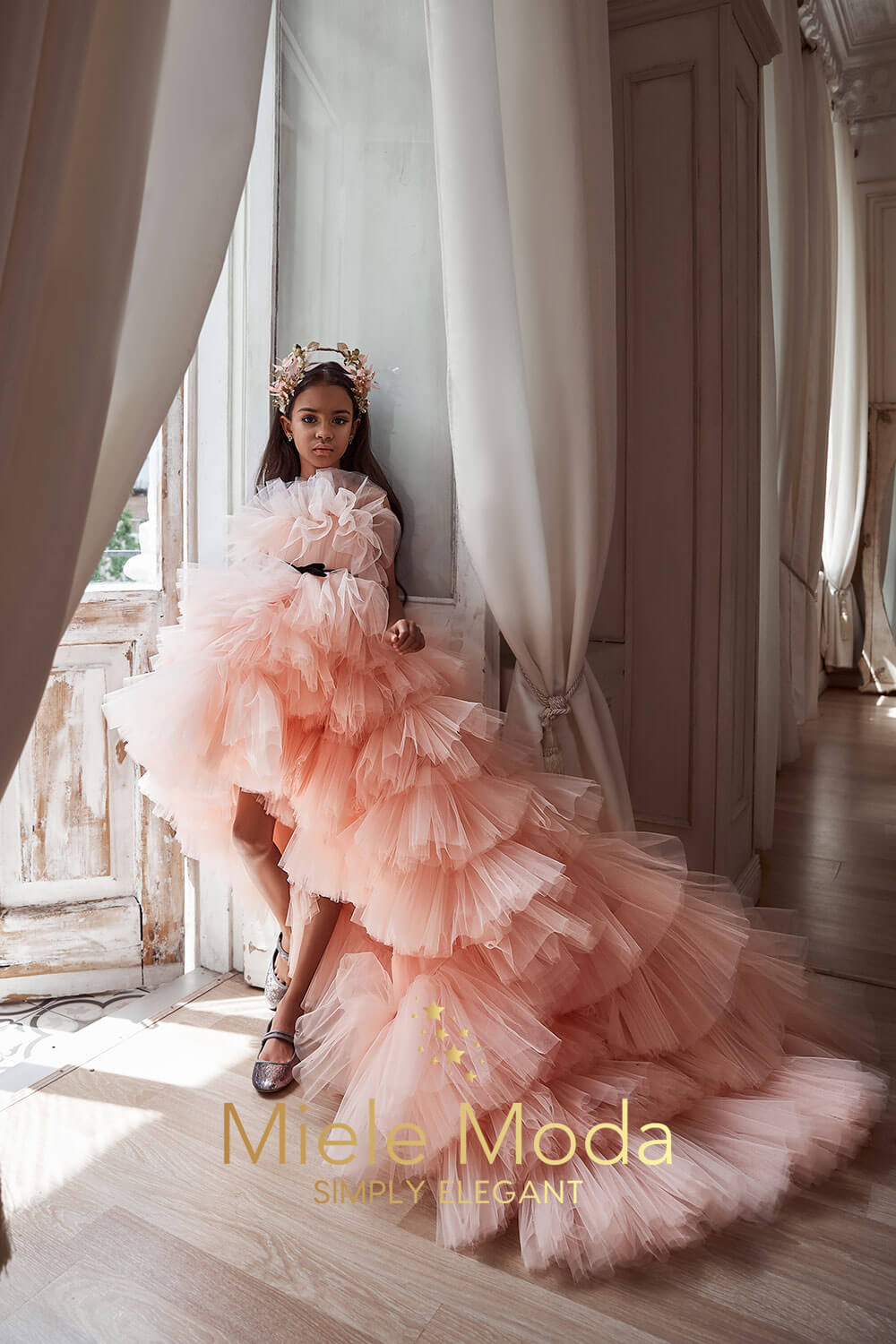 Young girl wearing a luxurious peach high-low ruffled tulle gown with a dramatic train, standing elegantly by a window framed with white curtains. She is accessorized with a floral crown and sparkly shoes.