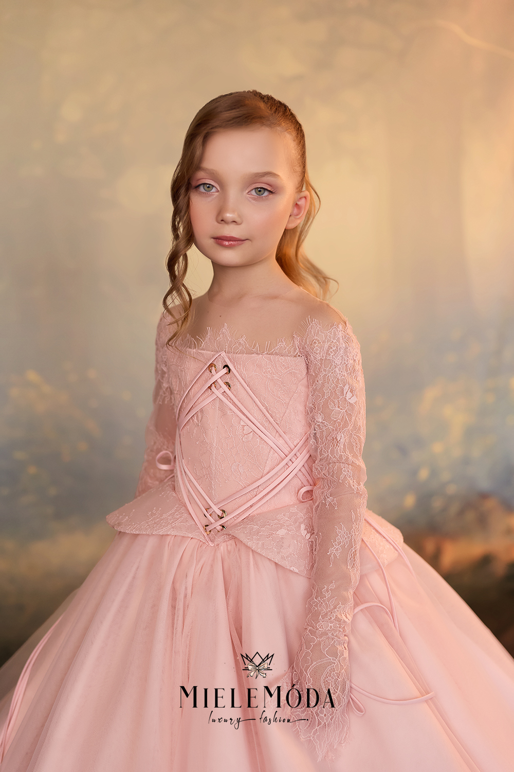 A young girl with light brown hair styled in braids, wearing a pale pink ballgown with lace long sleeves and a lace-up bodice, posed against a soft, painterly golden background.