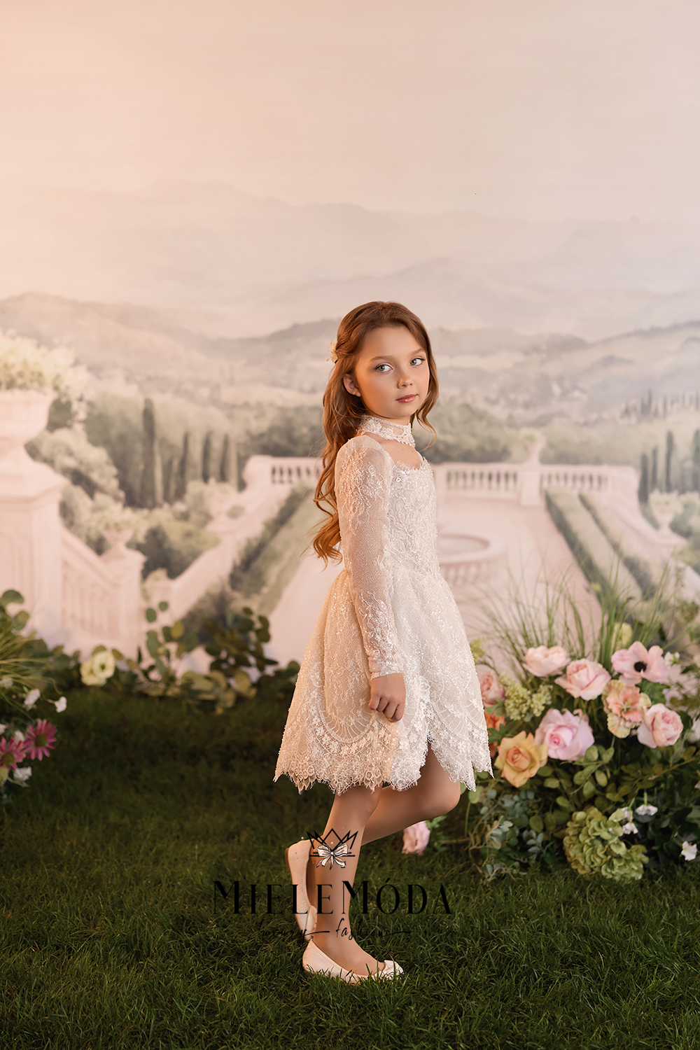 A young girl stands on a grassy lawn in front of a scenic garden backdrop, wearing a cream lace dress with long sleeves and a high neck, paired with beige flat shoes. The brand name 'MILLEMODA' is visible at the bottom.