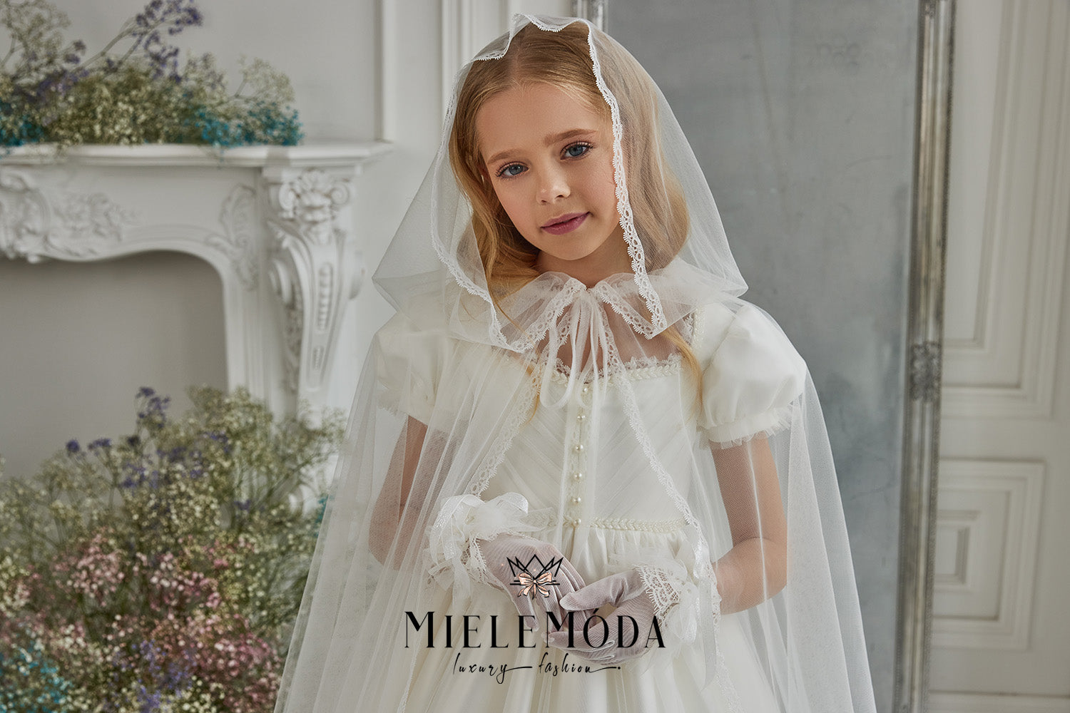 Young girl wearing a first communion dress, veil cape and gloves standing in front of a fireplace covered in flowers