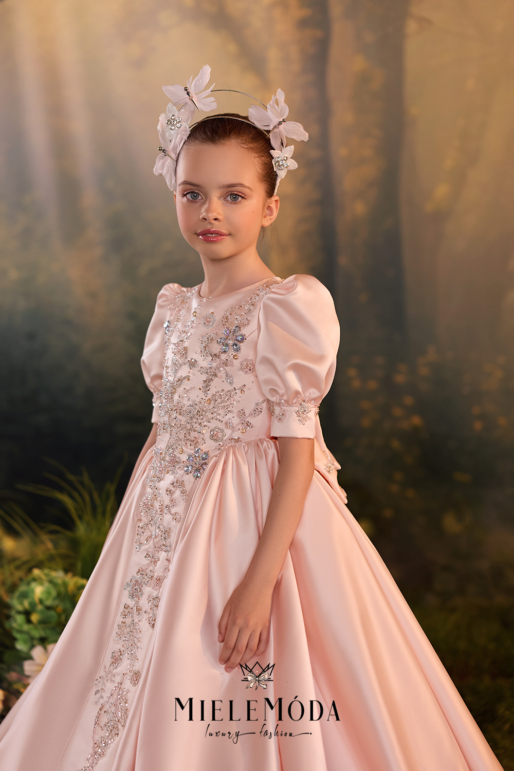 A young girl modeling a pale pink ballgown with intricate silver and blue beaded embroidery on the bodice and a matching floral headpiece, set against a soft-focus forest backdrop. The brand name 'MieleMóda' is visible at the bottom.