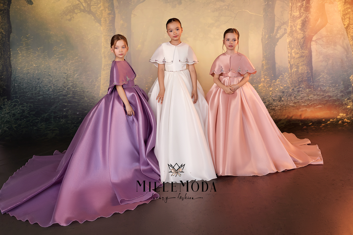 Three young girls modeling formal ball gowns in a studio setting with a forest backdrop. The girl on the left wears a lavender-purple gown, the center girl wears a white gown, and the girl on the right wears a light pink gown.