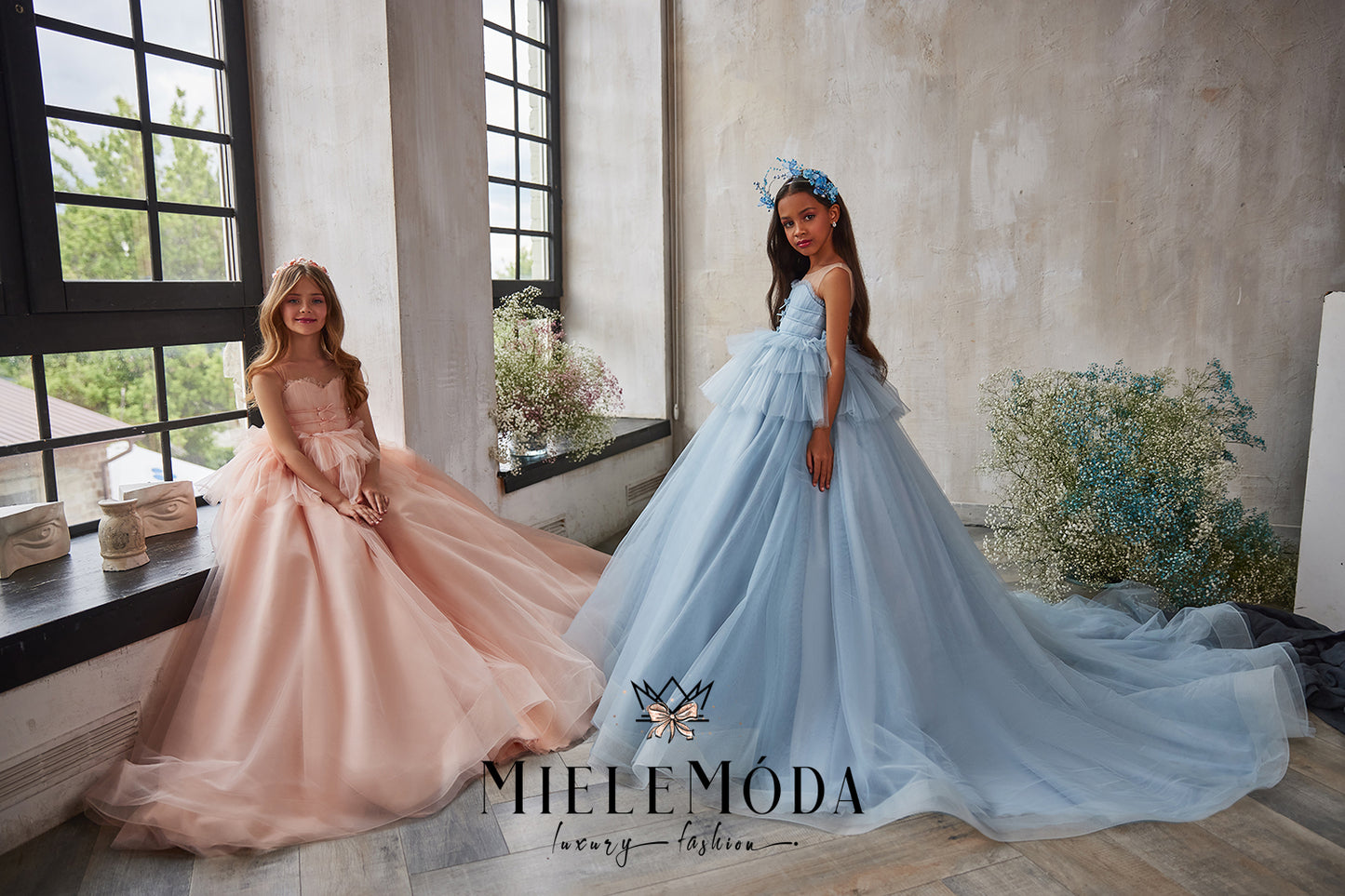 two gorgeous flower girls wearing pink and blue flower gowns sitting by window modeling for miele moda boutique