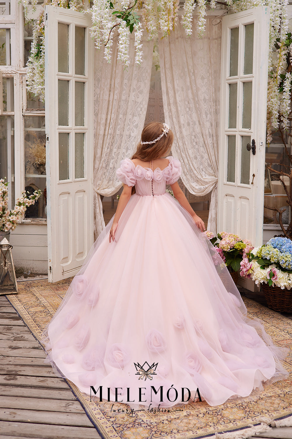 pretty flower girl wearing pink tulle dress with 3d flowers standing in front of open doors showing the back of the gown and headpiece