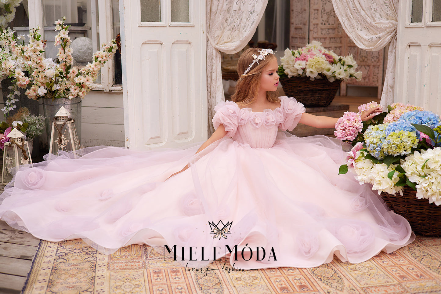 pretty flower girl wearing pink tulle dress with 3d flowers and pearl floral headpiece sitting in front of doors surrounded by flower baskets