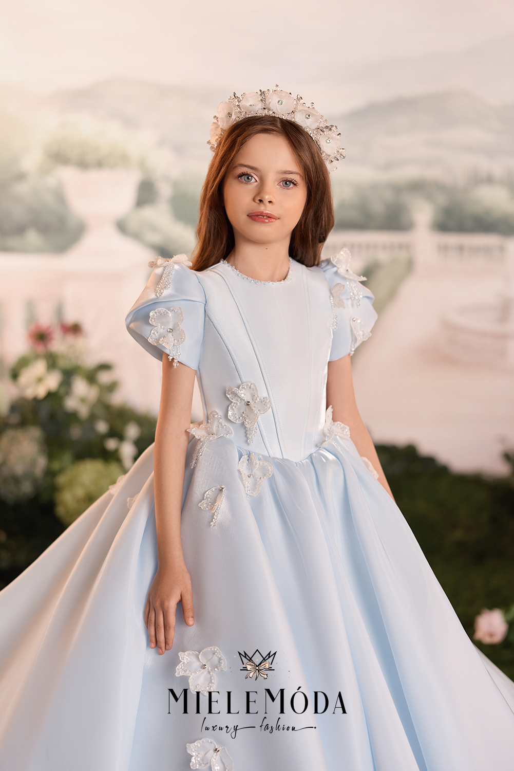 A young girl wearing a light blue ball gown with 3D floral appliqués and a matching floral crown, posed against a soft, blurred garden background.