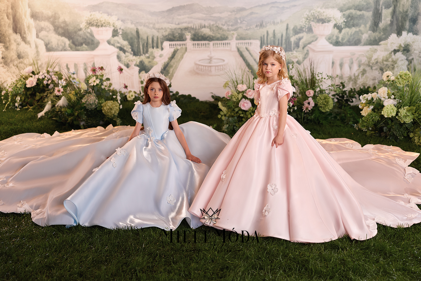 Two young girls wearing formal ball gowns pose on a green lawn in front of a large printed backdrop depicting a grand estate. The girl on the left wears a light blue gown with puffed sleeves and a long train, while the girl on the right wears a pale pink gown with ruffled sleeves, a fitted bodice, and a long train.