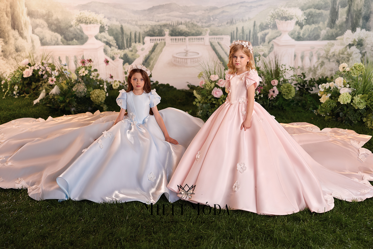 Two young girls wearing formal ball gowns pose on a green lawn in front of a large printed backdrop depicting a grand estate. The girl on the left wears a light blue gown with puffed sleeves and a long train, while the girl on the right wears a pale pink gown with ruffled sleeves, a fitted bodice, and a long train.