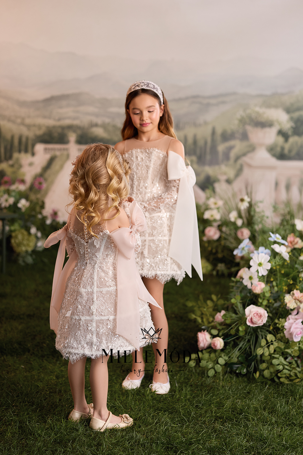 Two young girls modeling elegant off-the-shoulder lace dresses with white cape sleeves in a garden setting. The girl on the right wears a silver tiara, and the girl on the left has her hair in a blonde bun.