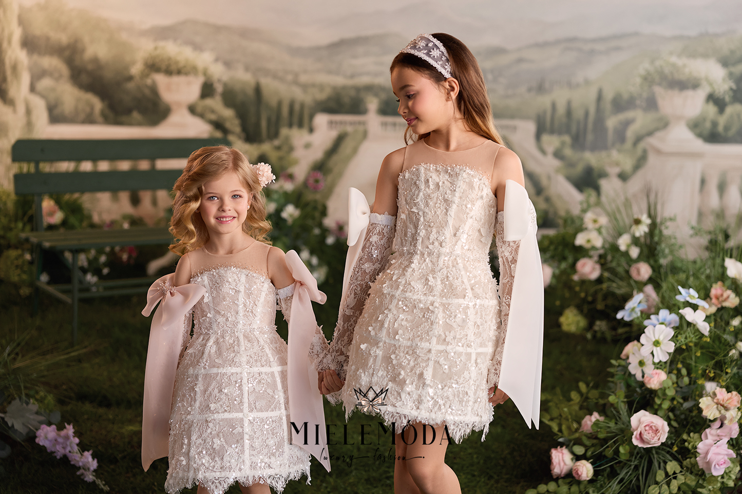 Two young girls wearing matching off-the-shoulder lace dresses with long sheer sleeves and draped white fabric accents, standing in a garden setting with a scenic backdrop. The brand name 'Mille Moda' is visible at the bottom center.
