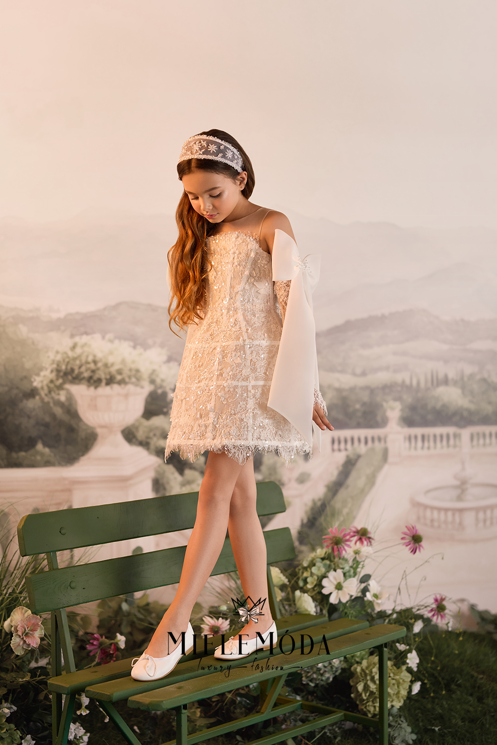 A young girl with long brown hair stands on a green bench wearing a cream lace off-the-shoulder dress, white flats, and a silver tiara, set against a soft, vintage-style landscape backdrop.