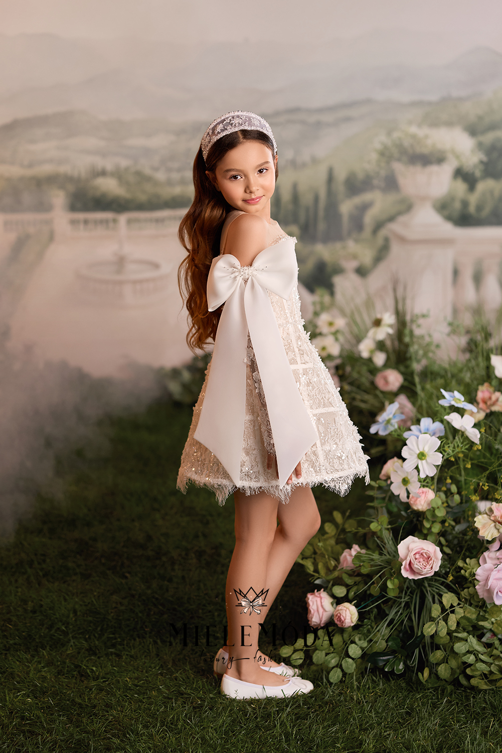 A young girl stands in a garden setting wearing a short cream lace dress with a large bow on the back, a matching lace headband, and white flats. The background is a soft, misty landscape with greenery and flowers.