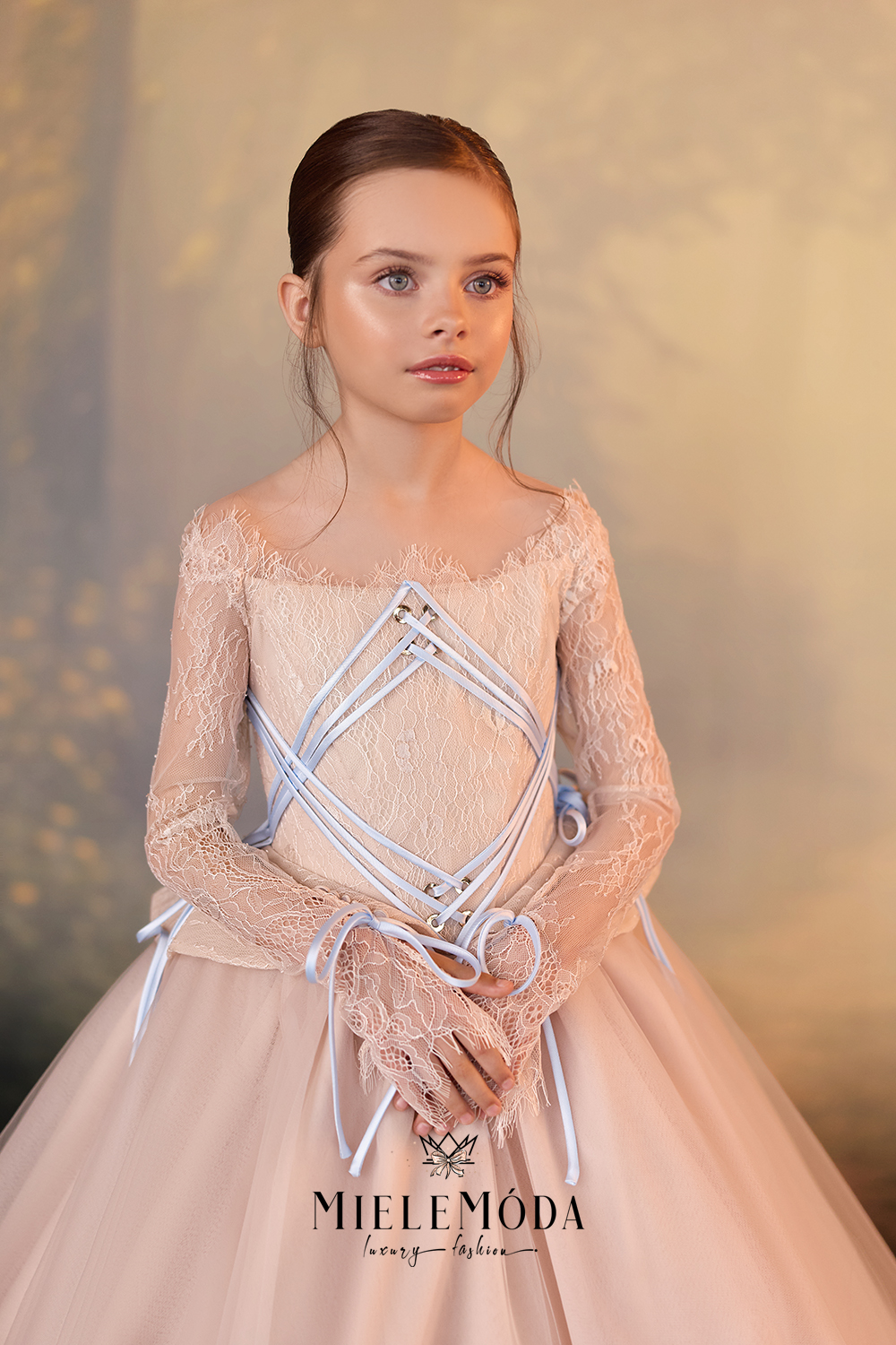 A young girl modeling a formal lace dress with a pale pink tulle skirt, sheer long sleeves, and a corset-style bodice with white lace-up details, posed against a soft beige background. The brand name MIELE MÓDA is visible at the bottom.