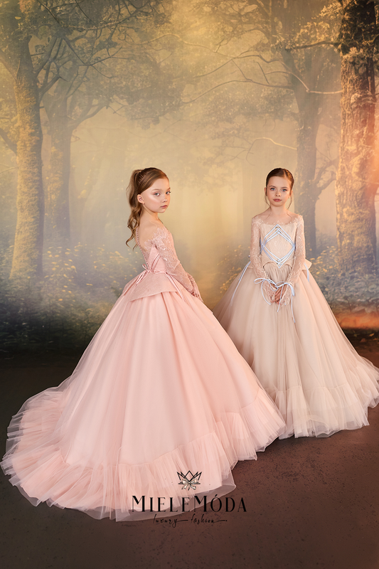 Two young girls wearing elegant ball gowns posed against a mystical, glowing forest backdrop. The girl on the left wears a voluminous light pink gown with a fitted bodice and full tulle skirt, while the girl on the right wears a pale blush-beige gown with a lace-up bodice and long sheer sleeves.