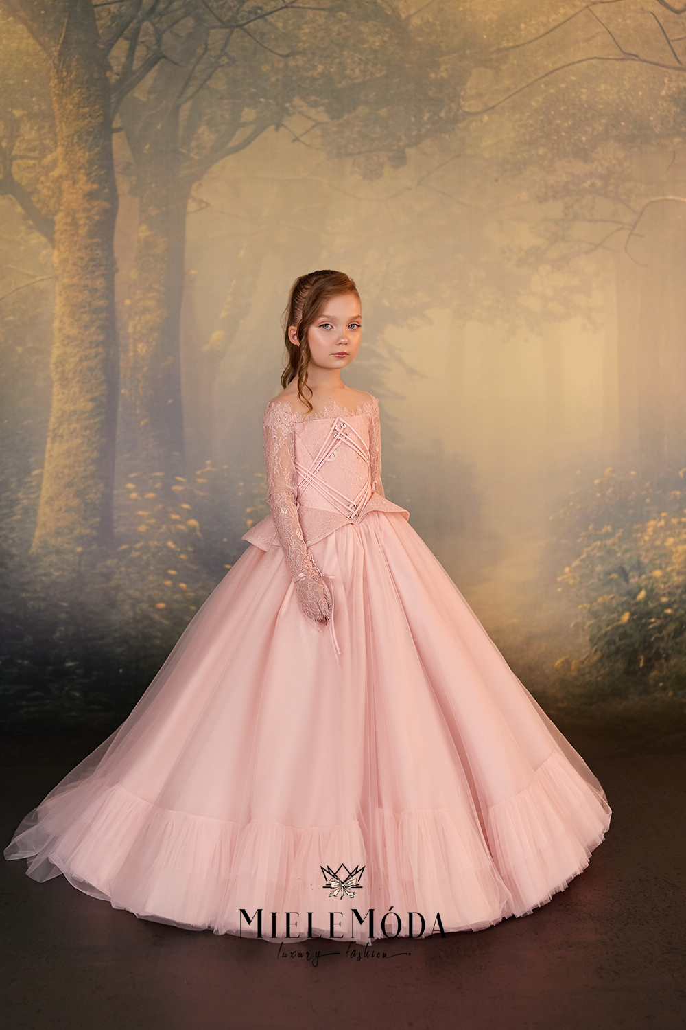 A young girl wearing a voluminous light pink ball gown stands in a misty, golden-toned forest scene. The dress has long sheer sleeves, a fitted bodice, and a full layered tulle skirt.