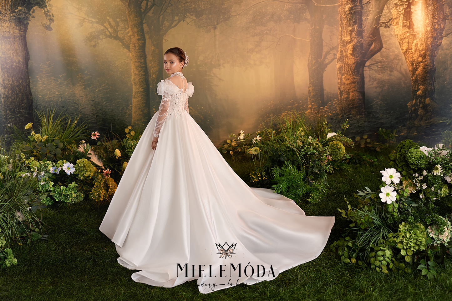 A woman wearing a voluminous white wedding gown stands in a forest-themed studio setting with misty trees, warm golden light, and floral arrangements around her.