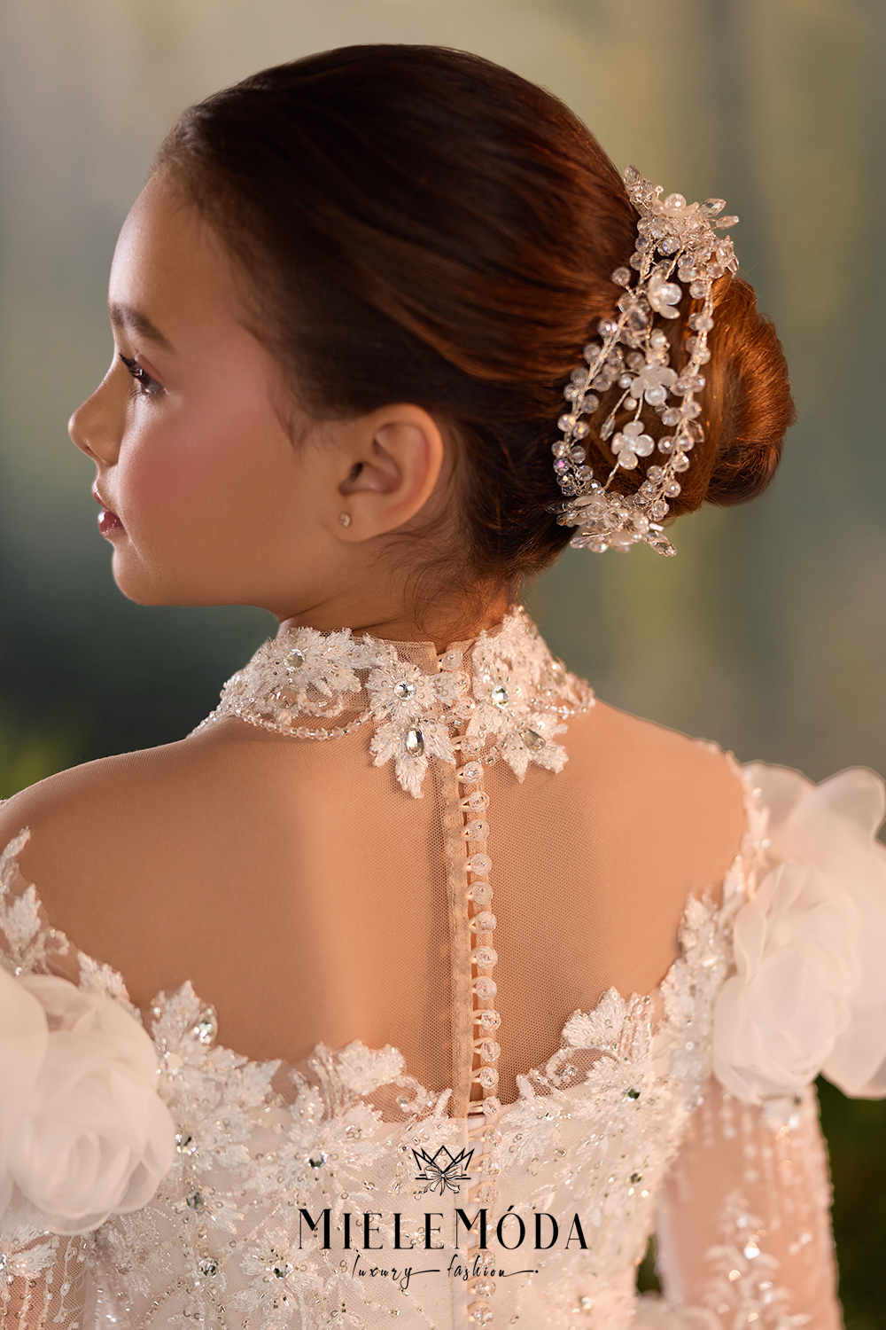 A young girl shown from the back wearing an ornate white lace bridal gown with floral details and a high embellished neckline, styled with a pearl and crystal hair accessory in her updo.