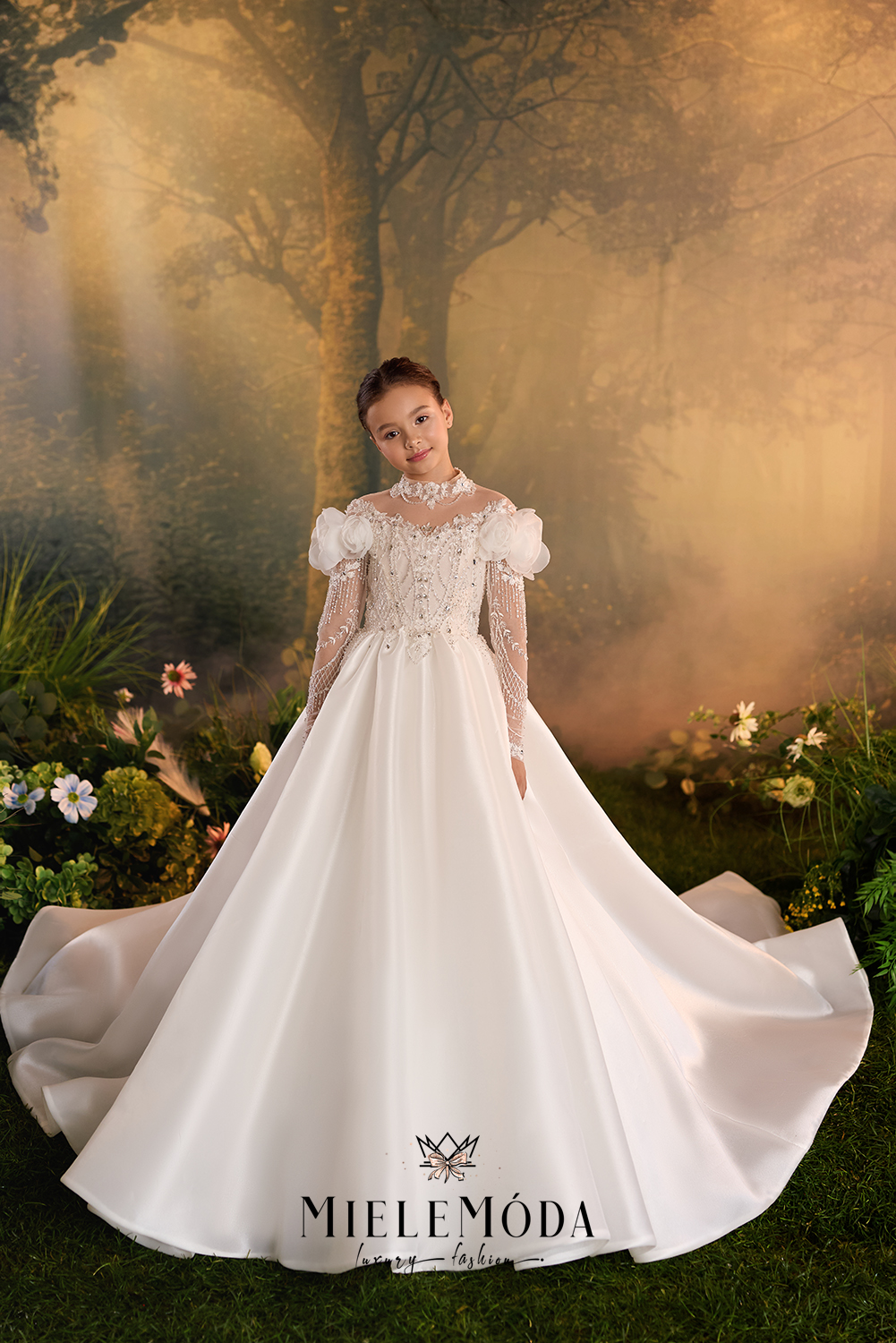 A young girl models a Miele Móda white ball gown with puffed sleeves, lace bodice, and a full satin skirt in a mystical, sunlit forest setting.