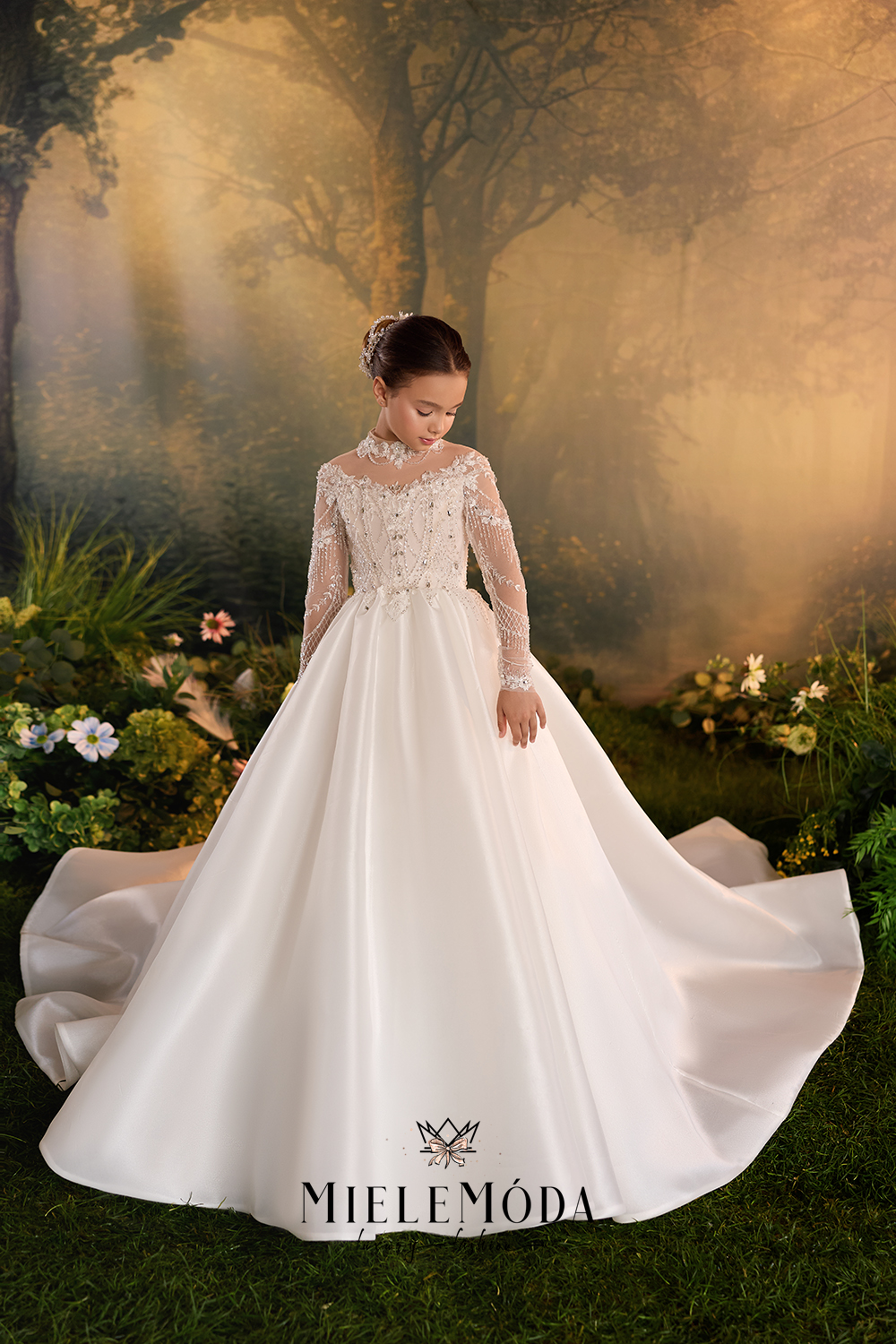 A young girl wearing a long-sleeved white ball gown with lace detailing and a full skirt, posed in front of a forest backdrop with the brand name MIELEMODA at the bottom.