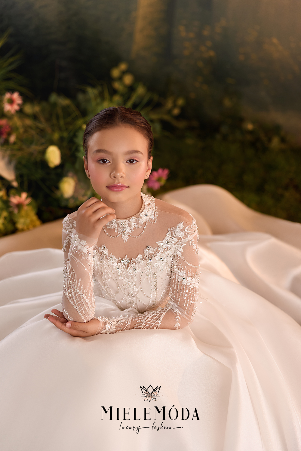 A young girl wearing a long-sleeved white lace wedding dress with a sheer embroidered bodice, posed in front of a soft-focus garden backdrop. The brand name 'MIELEMÓDA' is printed at the bottom.