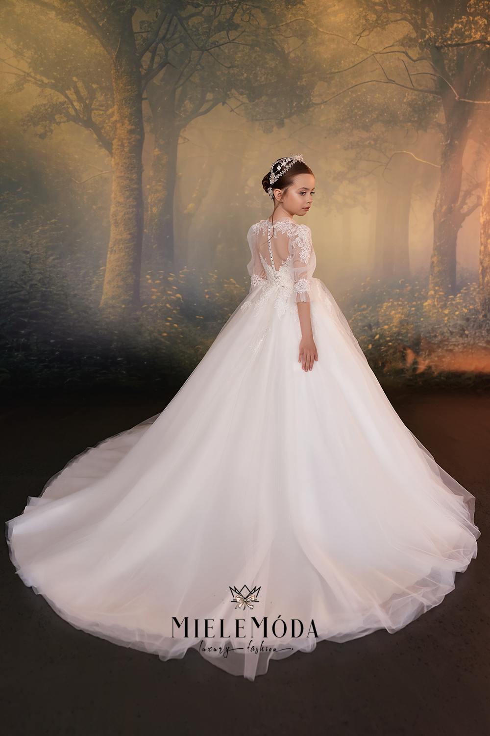 A young girl wearing a white wedding dress with lace long sleeves, a sheer back, and a full tulle skirt, posed in front of a misty forest backdrop. The brand name MIELEMODA is visible at the bottom.