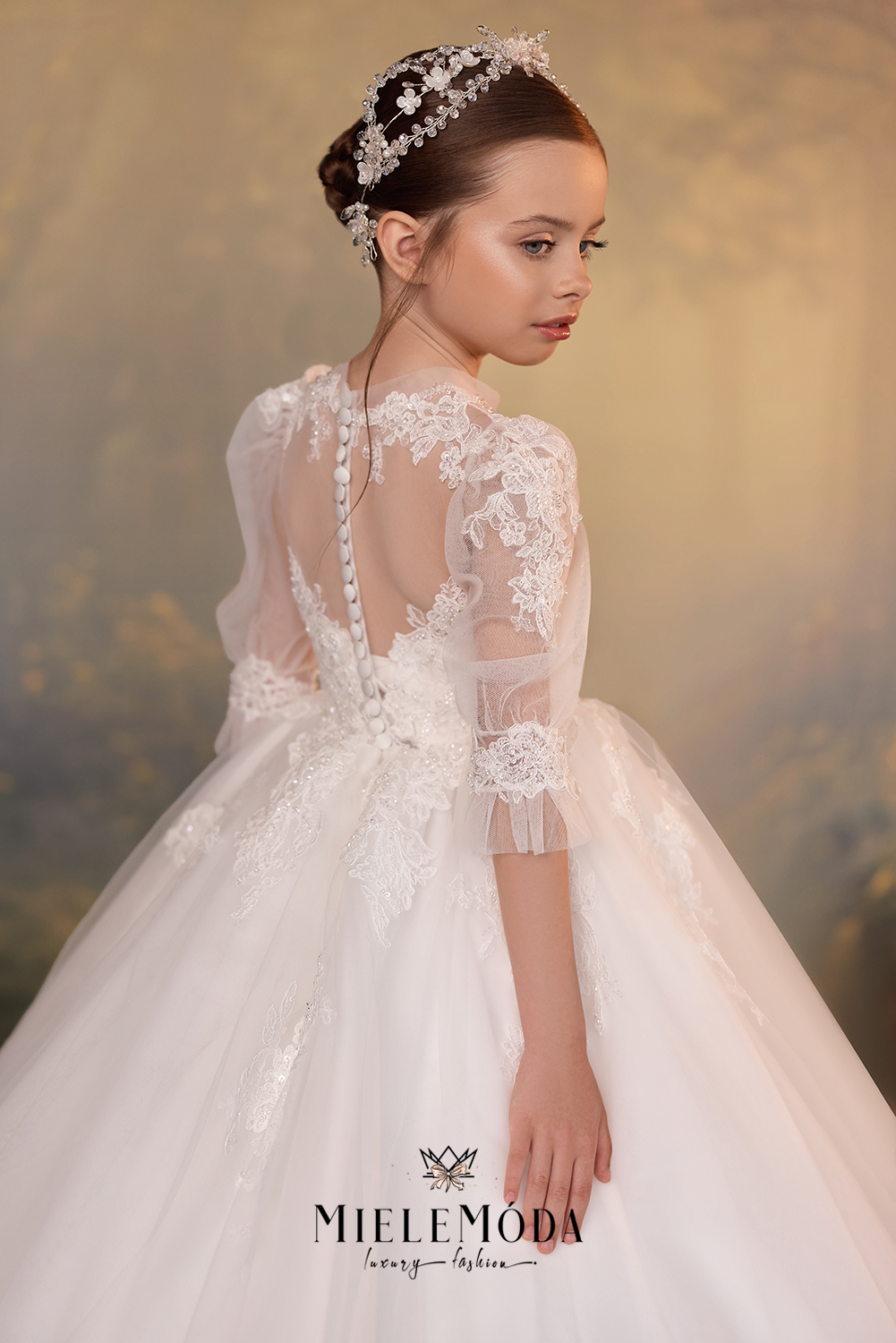 A young girl wearing a white lace and tulle formal dress with a sheer back, decorated with a pearl-like down the center, and wearing a jeweled headpiece. The brand logo 'MIELEMODA' is visible at the bottom.