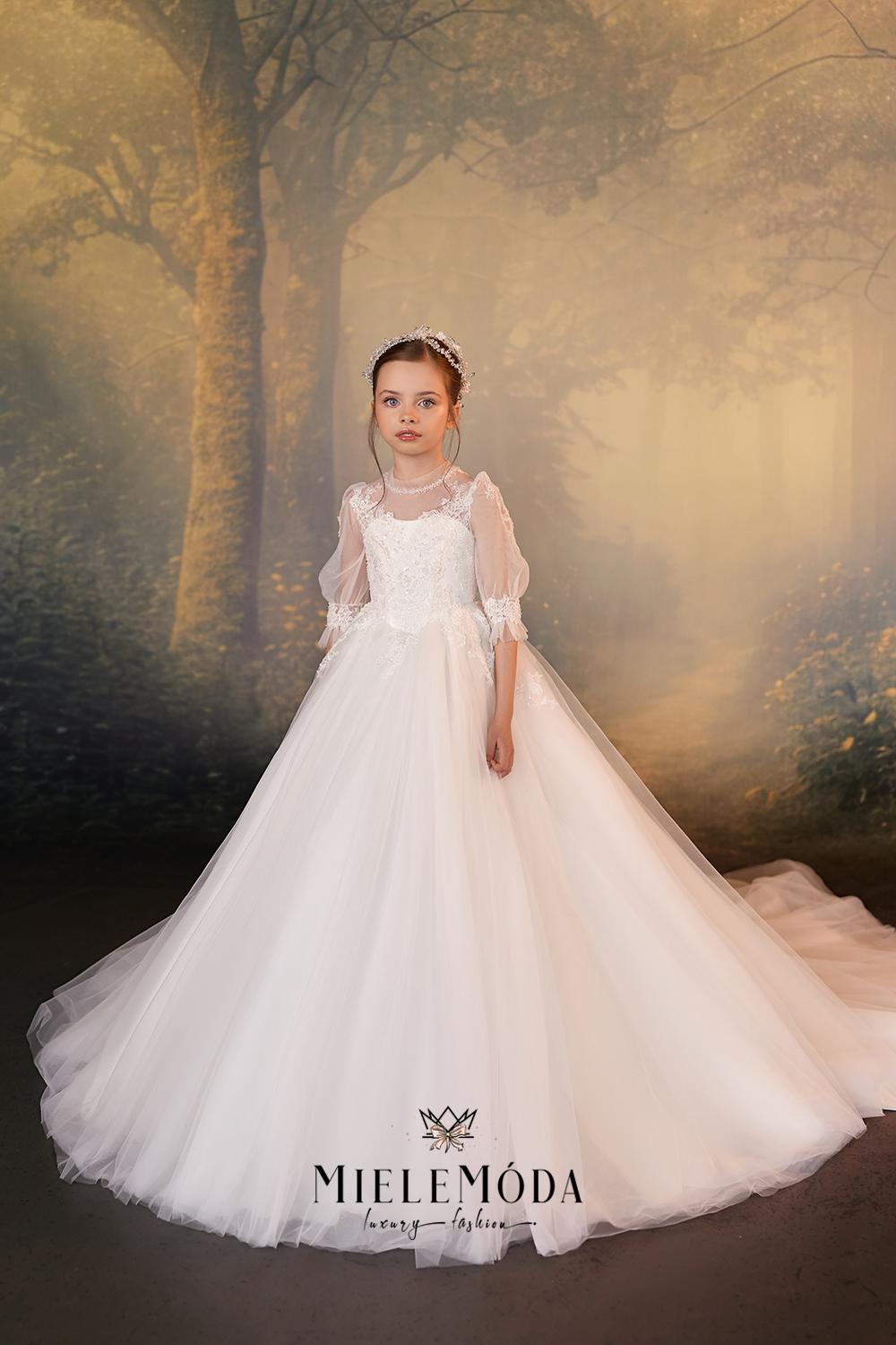 A young girl stands in a voluminous white formal dress with sheer puff sleeves and a full tulle skirt, posed against a misty forest backdrop. The brand name MIELEMODA is printed at the bottom of the dress.