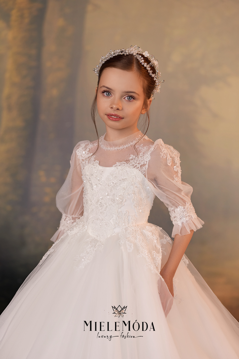 A young girl wearing a white lace and tulle ball gown with sheer puff sleeves, a matching lace collar, and a pearl tiara, posed against a warm, blurred studio backdrop. The brand name 'MIELEÓMÓDA' is printed at the bottom.