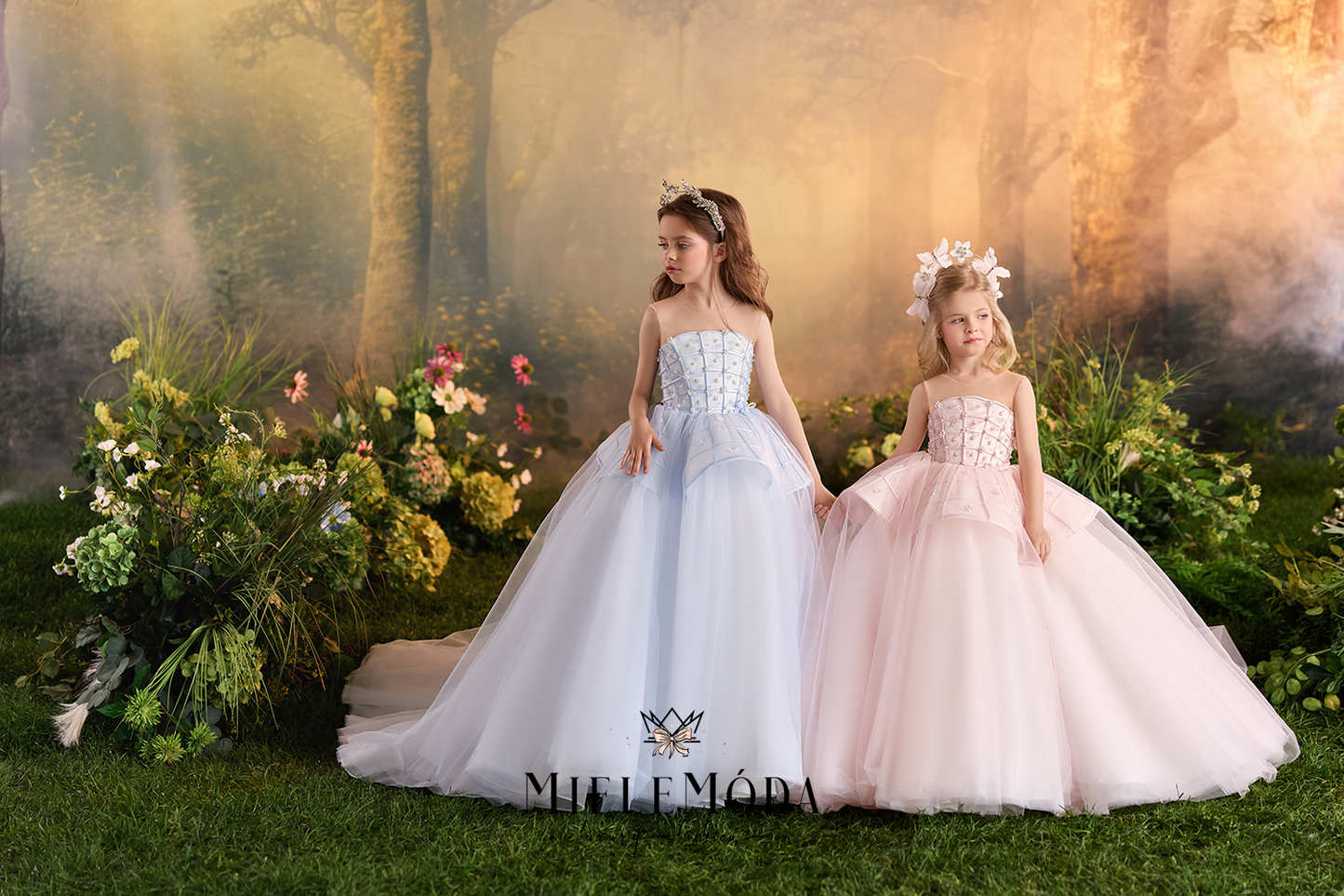 Two young girls in strapless tulle ballgowns stand in a fantasy forest setting. The girl on the left wears a light icy blue gown with a sheer embroidered bodice and full white tulle skirt.