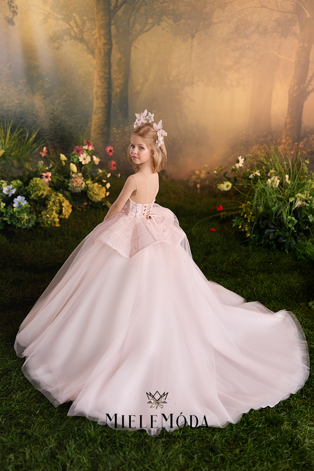 A young girl wearing a pale pink ball gown with a floral crown, posed in a magical, misty forest garden. The brand name 'MIELEMODA' is visible at the bottom of the image.