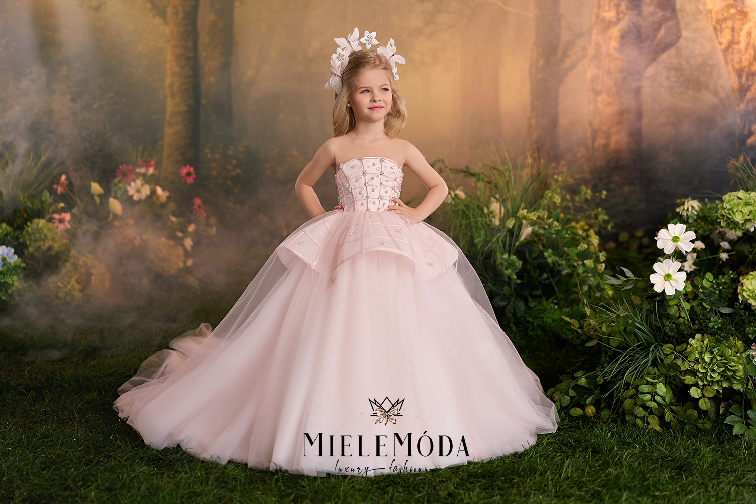A young girl stands in a fantasy forest setting wearing a strapless blush pink ball gown with a full tulle skirt and a matching floral headpiece.