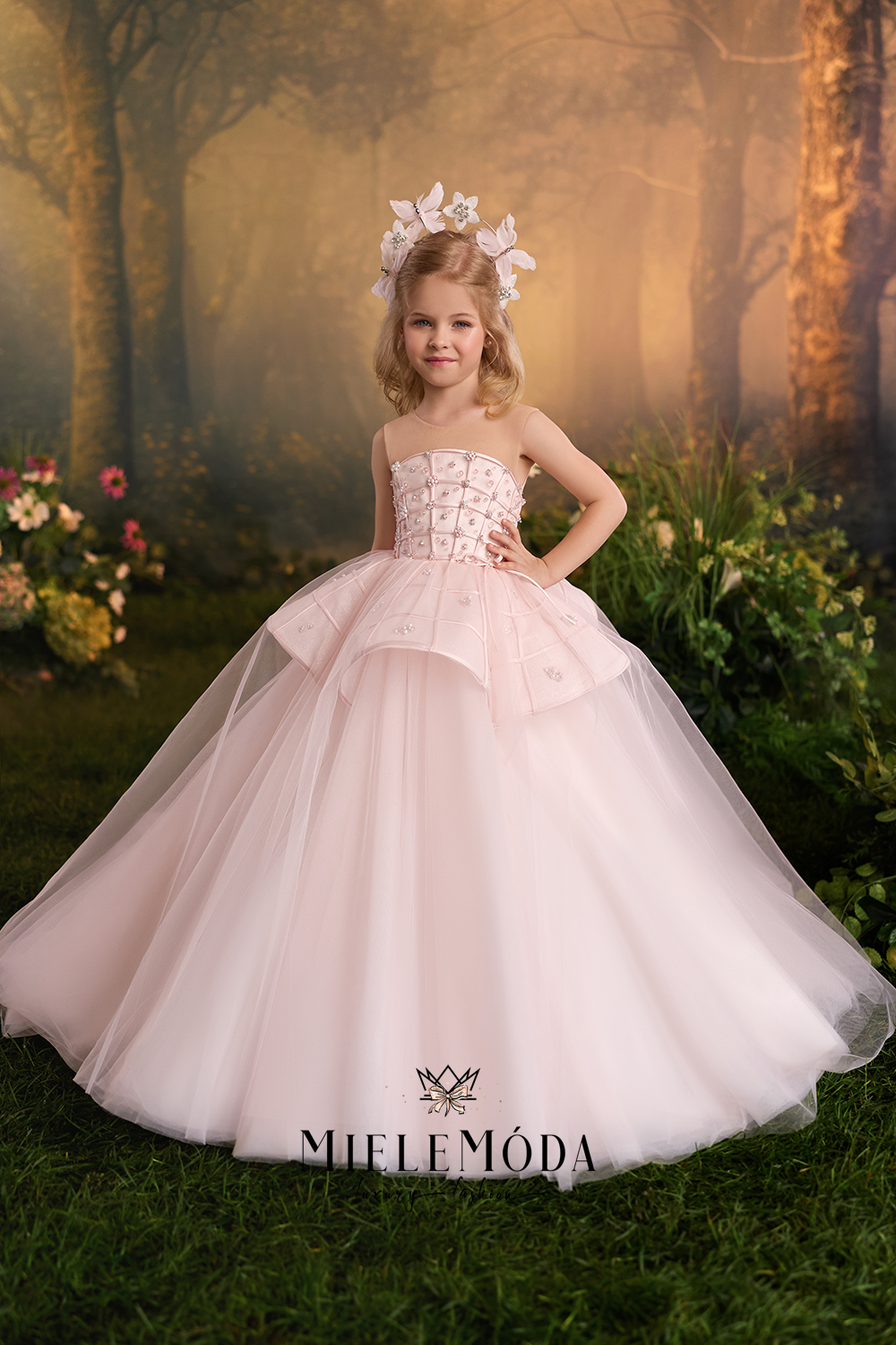 A young girl stands in a fantasy forest setting wearing a pale blush pink ball gown with a strapless bodice, layered tulle skirt, and a white floral headpiece.