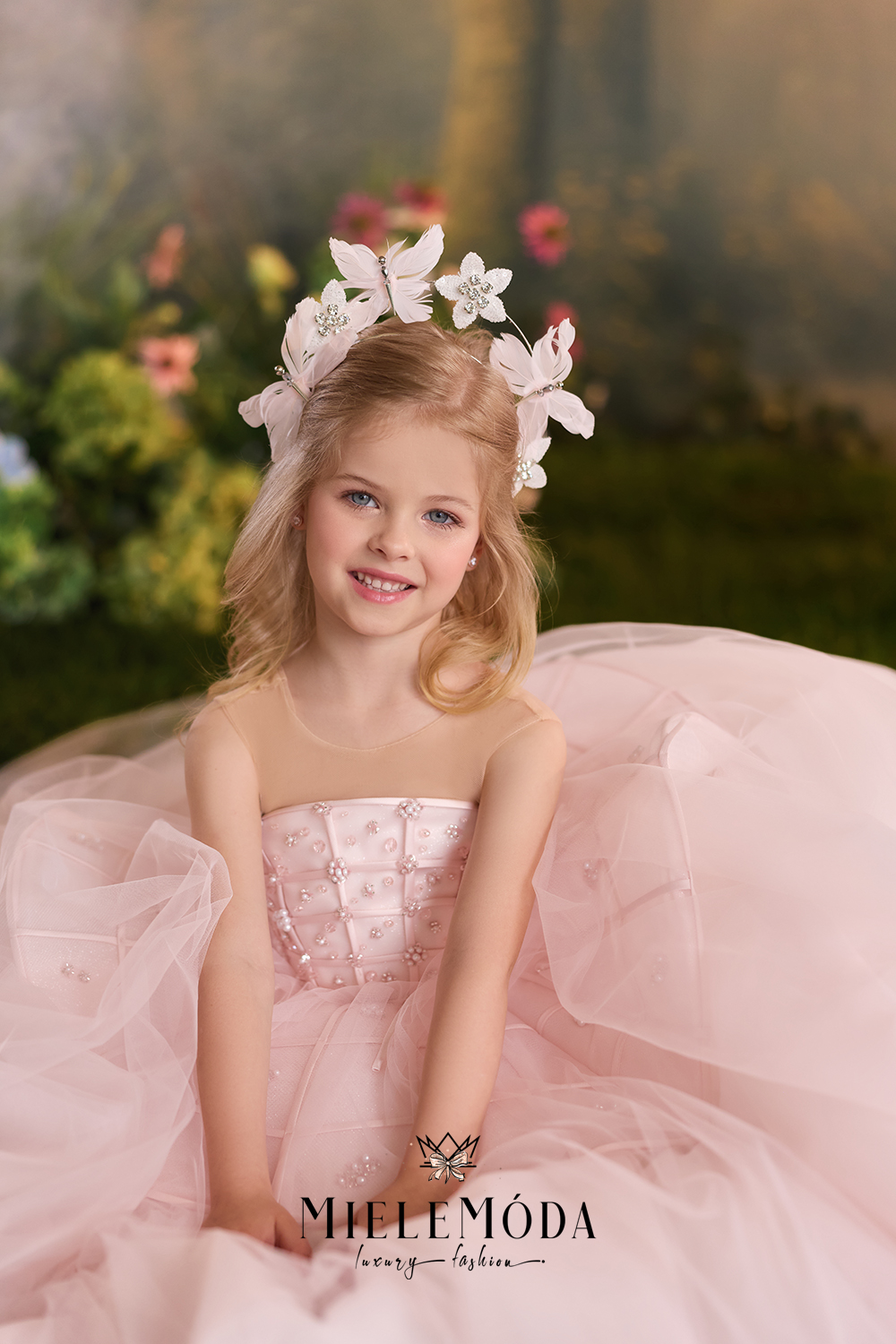 A young girl with blonde hair wearing a white floral crown sits in a pink tulle dress, smiling at the camera. The background is a soft-focus garden setting with green foliage and warm lighting.