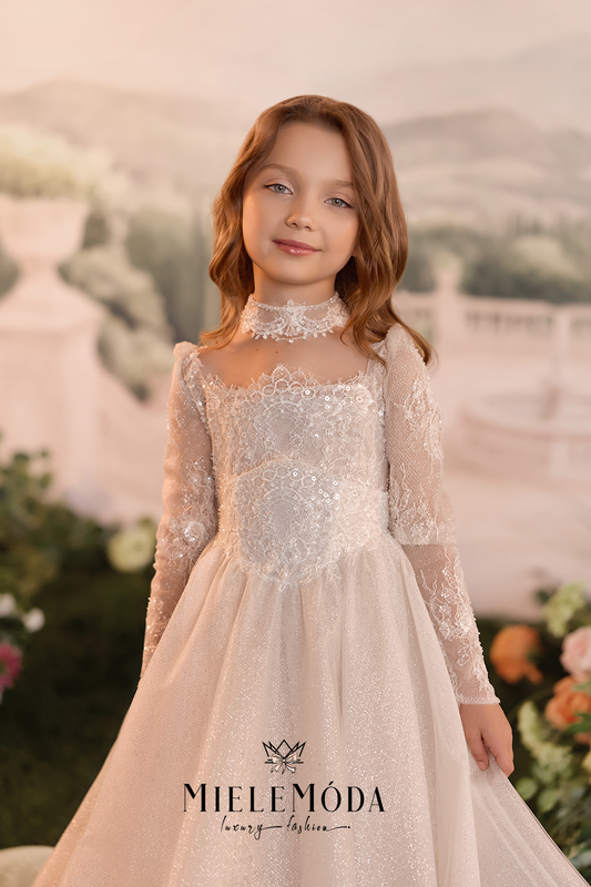 A young girl with wavy light brown hair wearing a long-sleeved ivory lace dress with a high lace collar and a sparkly tulle skirt, standing in front of a soft-focus garden backdrop.