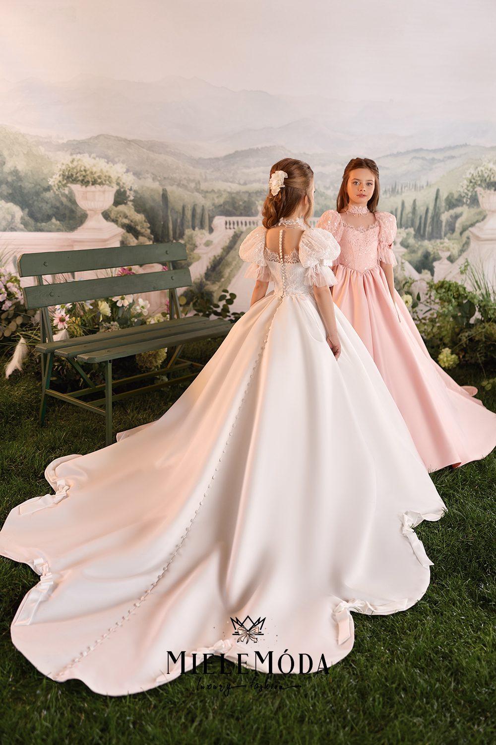 Two young girls modeling formal ballgowns in an outdoor garden setting with a painted backdrop of hills and a bench. The girl in the foreground wears a large off-white/ivory gown with puffed sleeves and a long train, while the girl behind wears a light pink gown with similar styling.
