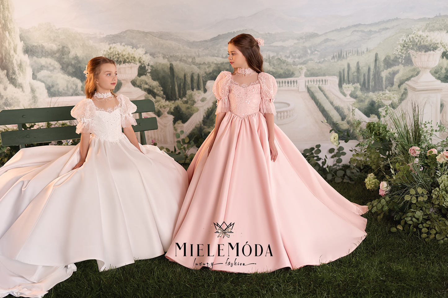 Two young girls wearing elegant ballgowns posed in front of a scenic garden backdrop. The girl on the left wears a white gown with puffed sleeves, and the girl on the right wears a dusty pink gown with similar puff sleeves.