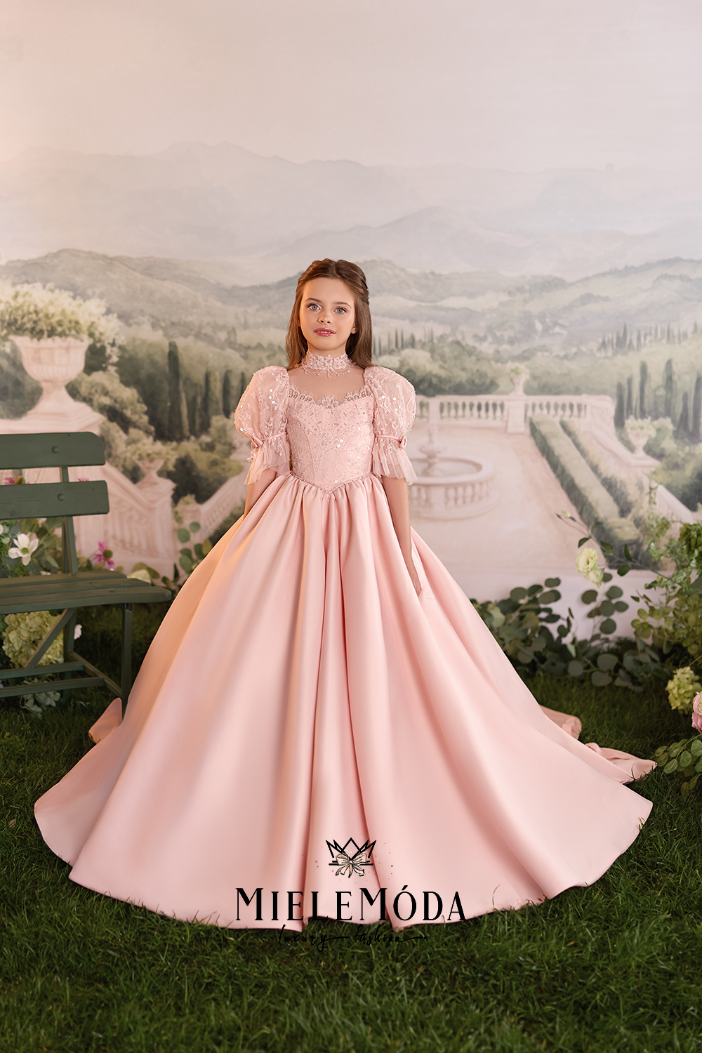 A young girl wearing a pale pink ball gown with puff sleeves and a lace bodice, standing in front of a scenic garden backdrop. The brand name MIELEMÓDA is displayed at the bottom.
