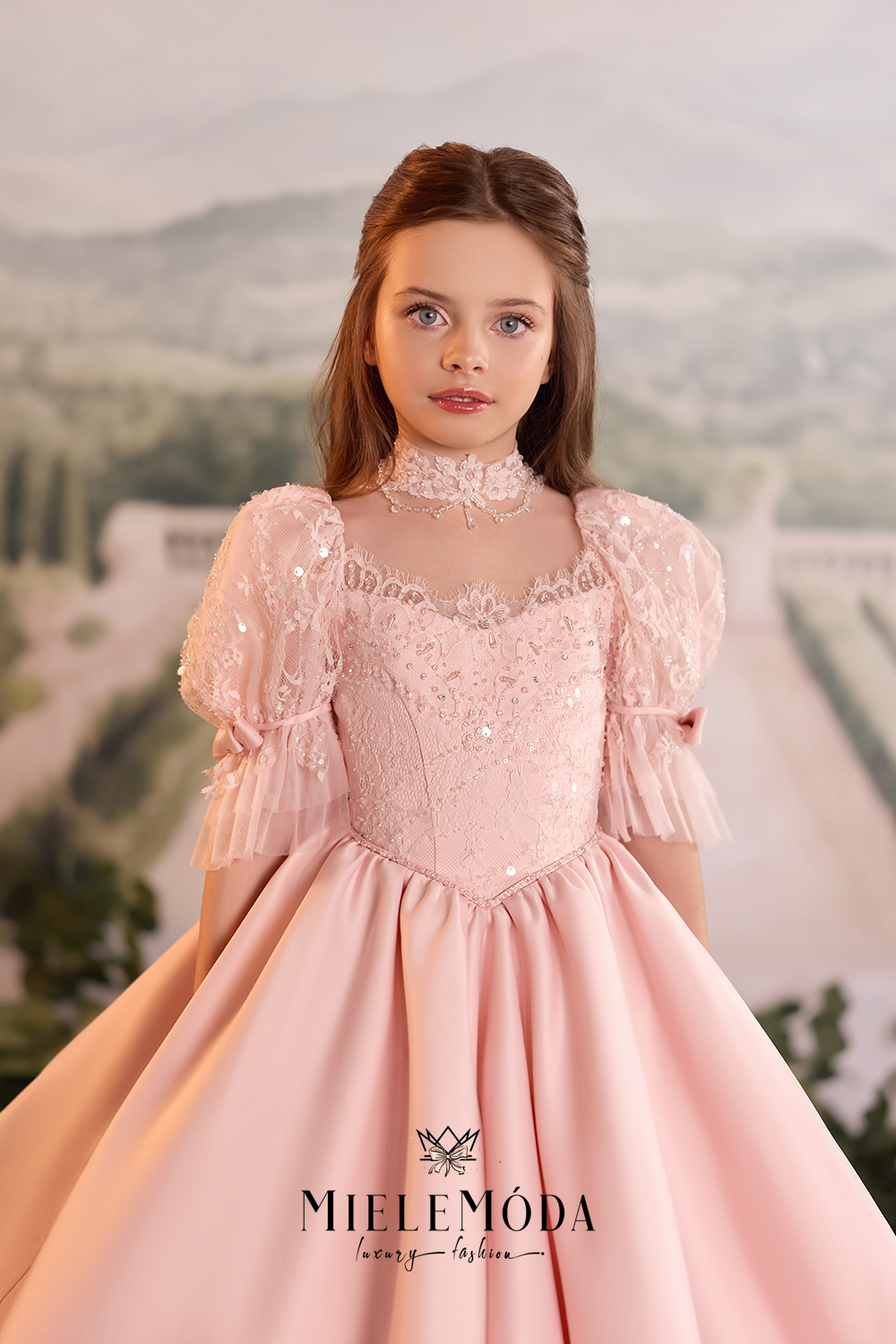 A young girl wearing a pale pink ballgown with a high lace collar, puffed sheer sleeves, and a full skirt, posed against a soft scenic backdrop. The brand name MIELEMÓDA is visible at the bottom of the dress.