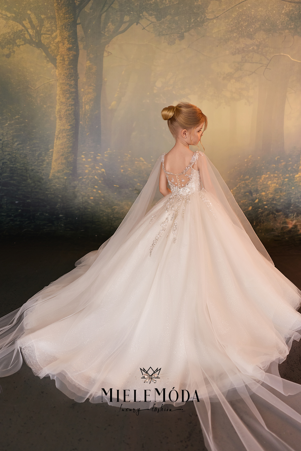 A young girl with her hair in a bun is shown from behind wearing a voluminous white tulle wedding dress with a long sheer veil, set against a misty forest backdrop. The brand name 'NIELEMODA' is visible at the bottom.
