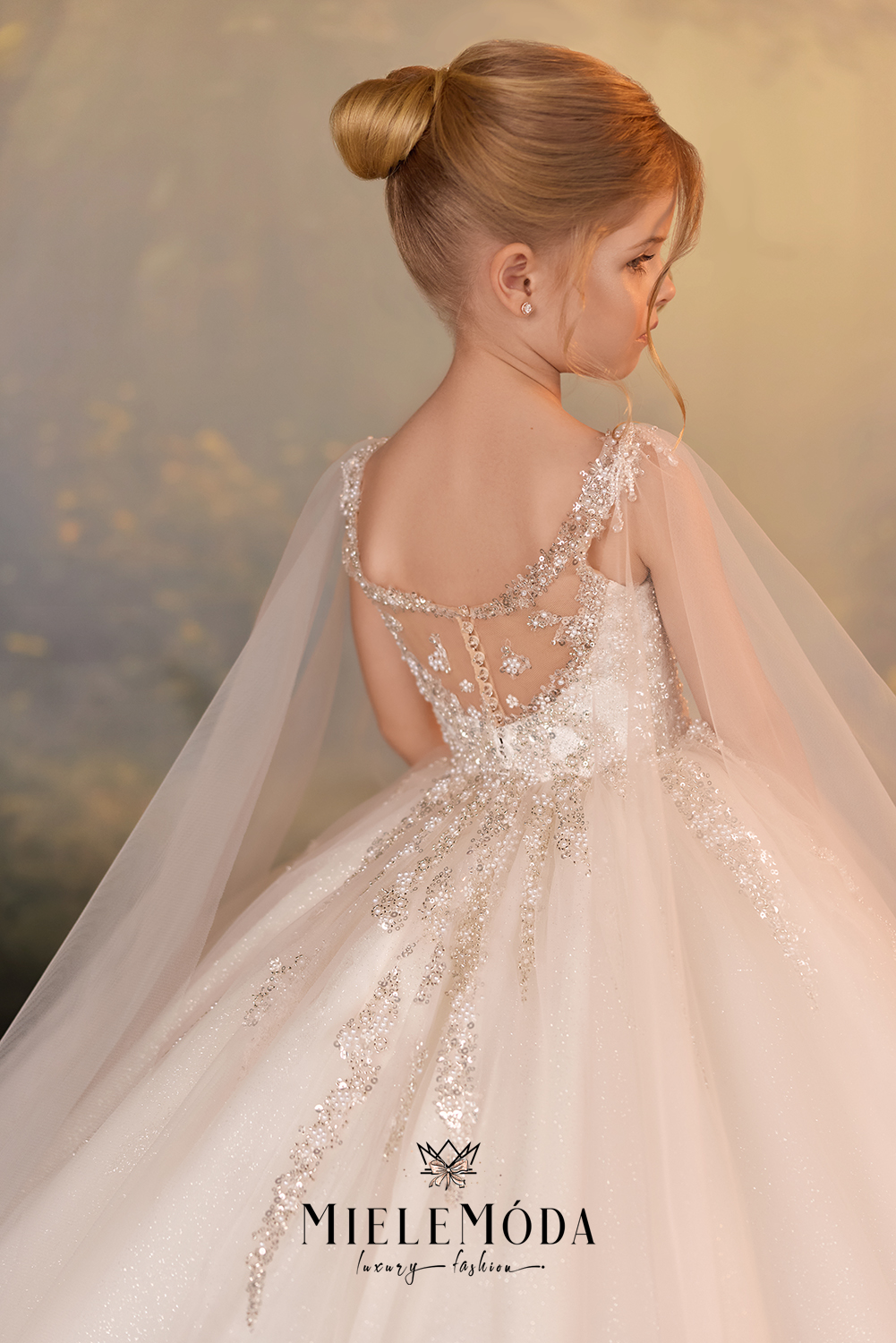 A young girl shown from the back wearing a Miele Móda ball gown with a sheer lace-up back, embellished beading, and a flowing tulle skirt with a sheer veil. The background is a soft, warm, blurred outdoor scene.