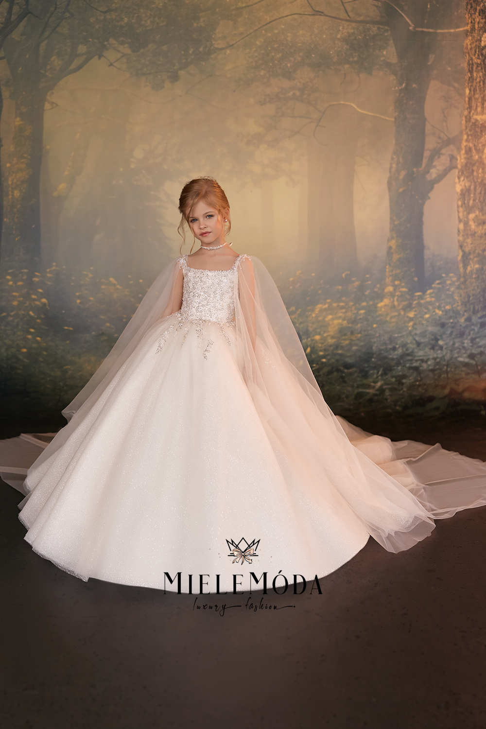 A young girl with red hair wearing a white ball gown with a sheer tulle cape, standing in front of a misty forest backdrop. The brand name 'MIELEMÓDA' is visible at the bottom.