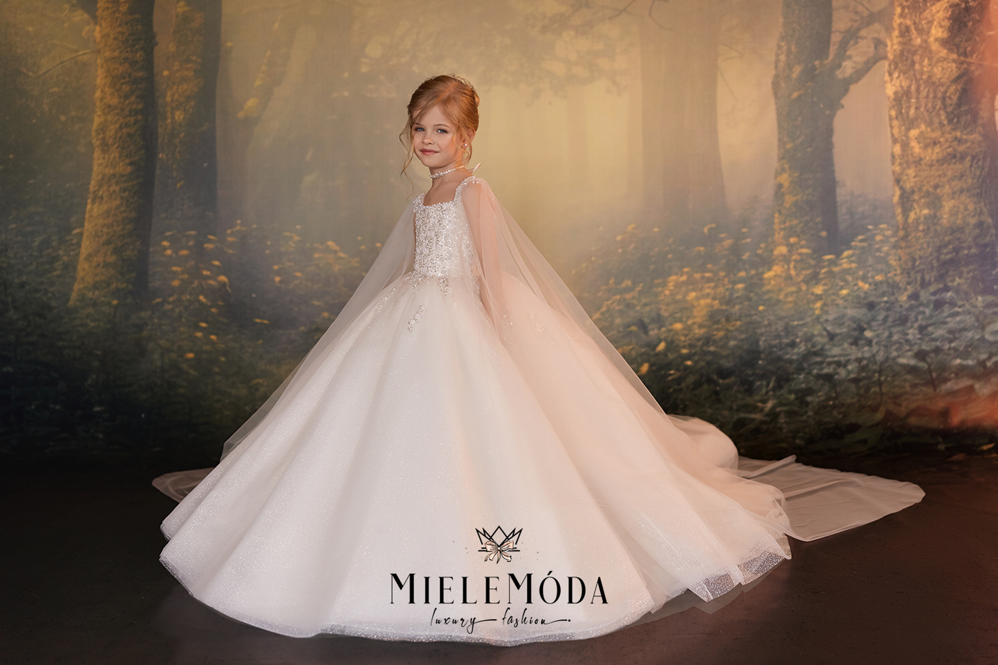 A young girl wearing a voluminous white tulle flower girl dress with a sheer veil, standing in front of a misty forest backdrop. The brand name 'MieleMóda' is printed on the dress.