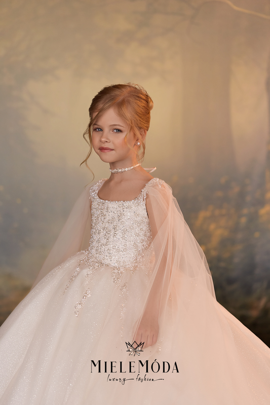 A young girl with styled light brown hair wears a formal white and pale pink ballgown with a sheer tulle cape and a pearl choker, posed against a soft, warm-toned forest backdrop. The brand name 'MIELEMÓDA' is visible at the bottom.