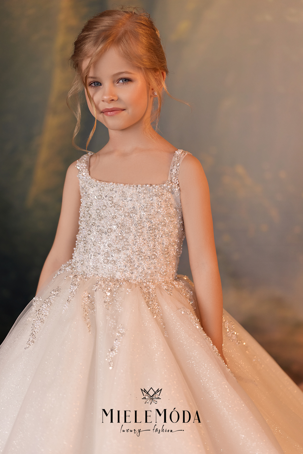 A young girl wearing a sparkling champagne-colored ball gown with intricate beadwork and a voluminous tulle skirt, posed against a soft, warm-toned studio backdrop. The brand name 'MIELEMÓDA' is visible at the bottom of the dress.