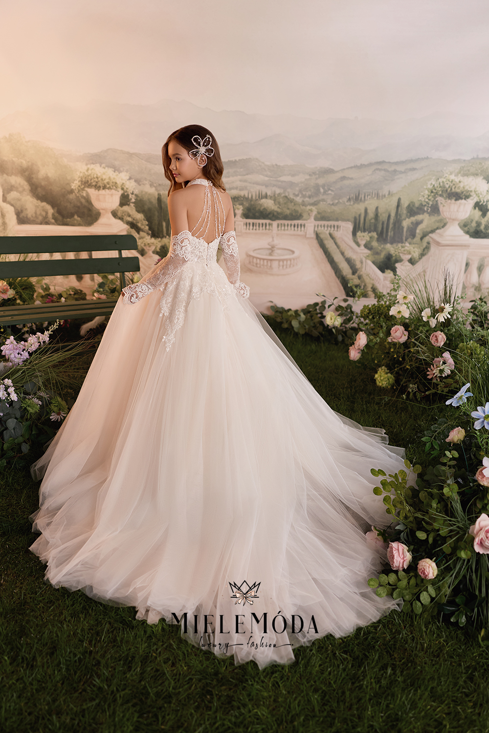 A bride in a voluminous off-white tulle wedding gown with lace back detailing and long sleeves stands on grass in front of a scenic garden backdrop with flowers and a bench. The brand name MIFLEMODA is visible at the bottom.