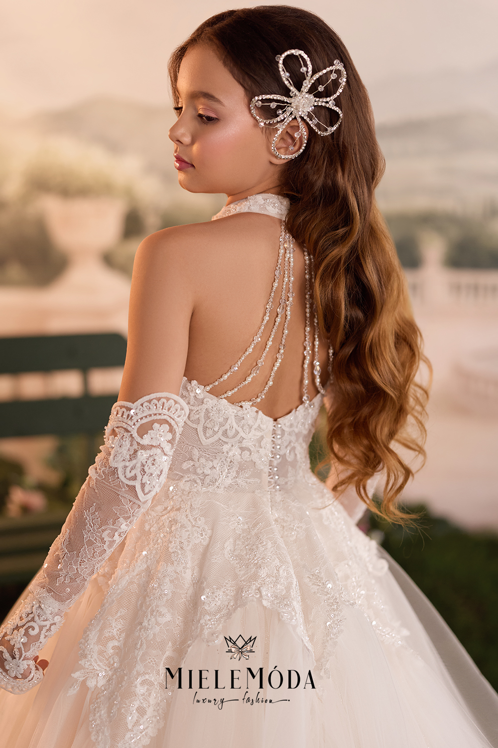 A young girl wearing a white lace bridal gown with a beaded halter back, off-the-shoulder lace sleeves, and a sparkling hair accessory, photographed outdoors with a soft blurred background. The brand name MIELEMODA is visible at the bottom.