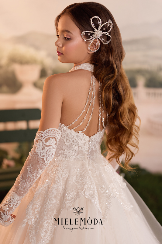 A young girl wearing a white lace bridal gown with a beaded halter back, off-the-shoulder lace sleeves, and a sparkling hair accessory, photographed outdoors with a soft blurred background. The brand name MIELEMODA is visible at the bottom.