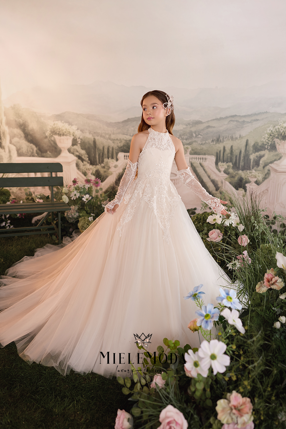 A young girl wearing a white lace and tulle ball gown stands in a garden-like setting with flowers, a bench, and a scenic painted backdrop. The brand name Mieleflood is visible at the bottom.