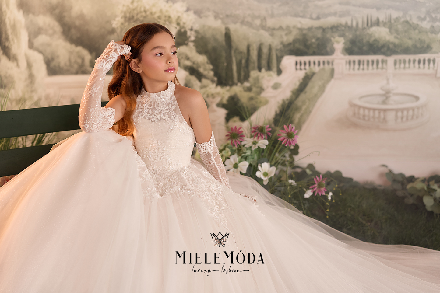 A young girl wearing a white lace and tulle ball gown with long sheer sleeves, posed in front of a scenic garden backdrop. The brand name 'Miele Möda' is visible at the bottom.