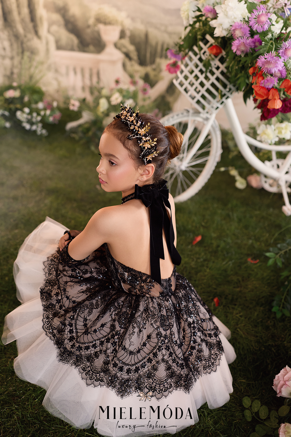 A young girl wearing a black lace and tulle flower girl dress with a black ribbon back bow, kneeling in a garden setting with a white decorative bicycle and flowers in the background. The brand name MieleModa is visible at the bottom.